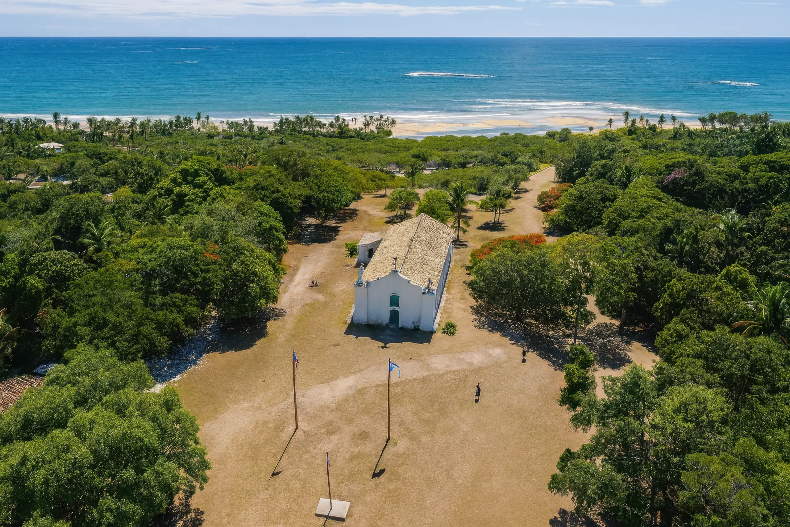 Vista aérea do Quadrado de Trancoso, com a igrejinha branca ao fundo e o mar azul no horizonte.